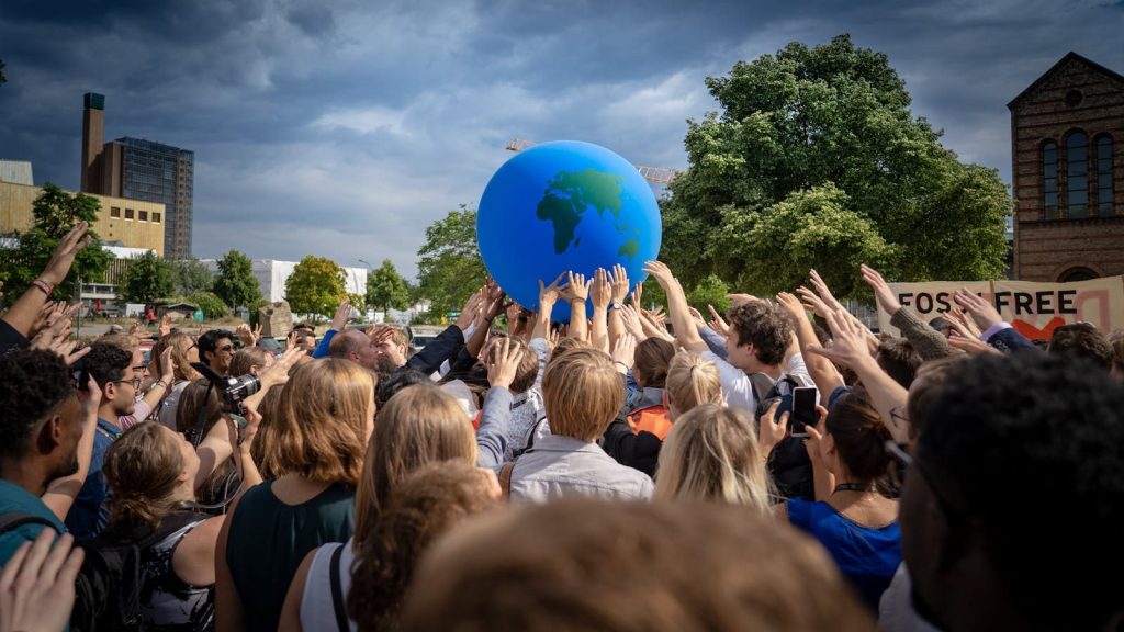 A diverse crowd participates in a climate protest in Berlin, Germany, raising a large Earth ball symbolizing environmental unity.