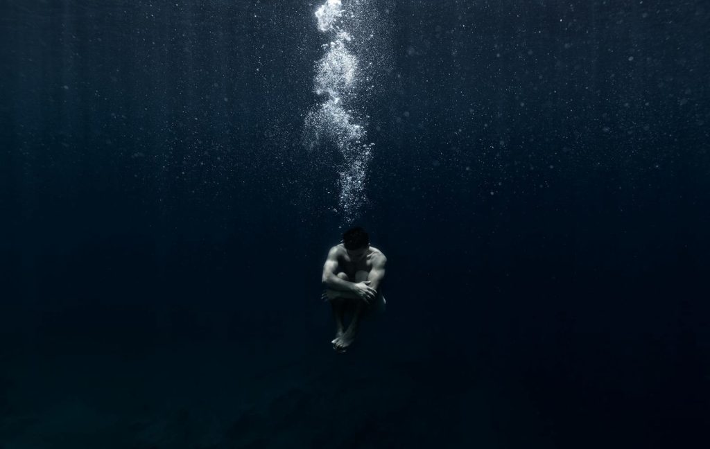 A solitary man floats submerged underwater, releasing air bubbles into the deep, dark ocean.