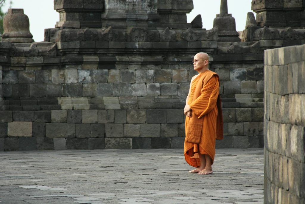 A Buddhist monk stands in orange robes at an ancient stone temple, depicting peace and spirituality.