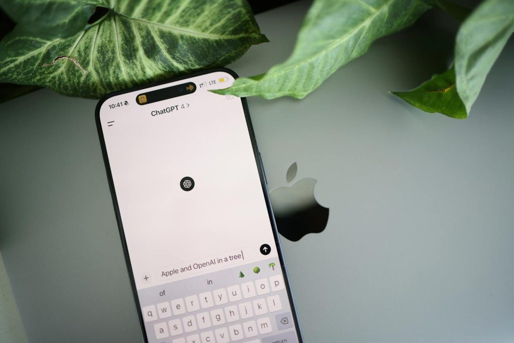 A smartphone shows a ChatGPT interface placed on an Apple laptop in a leafy environment.