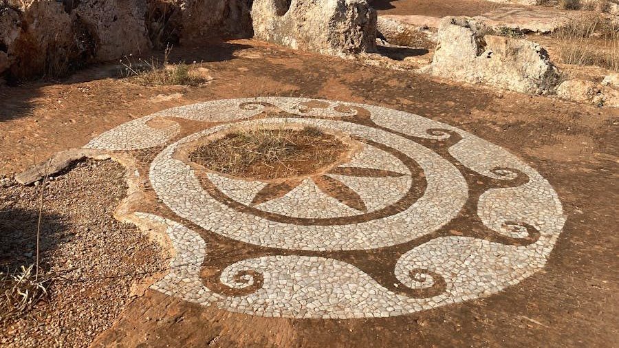 Explore the ancient mosaic patterns set against the rugged landscape of Vathia, Greece, under a clear blue sky.