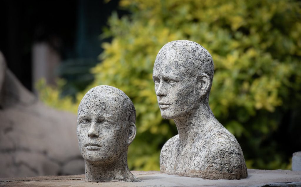 Two lifelike stone sculptures of faces against a leafy backdrop, showcasing artistic craftsmanship.