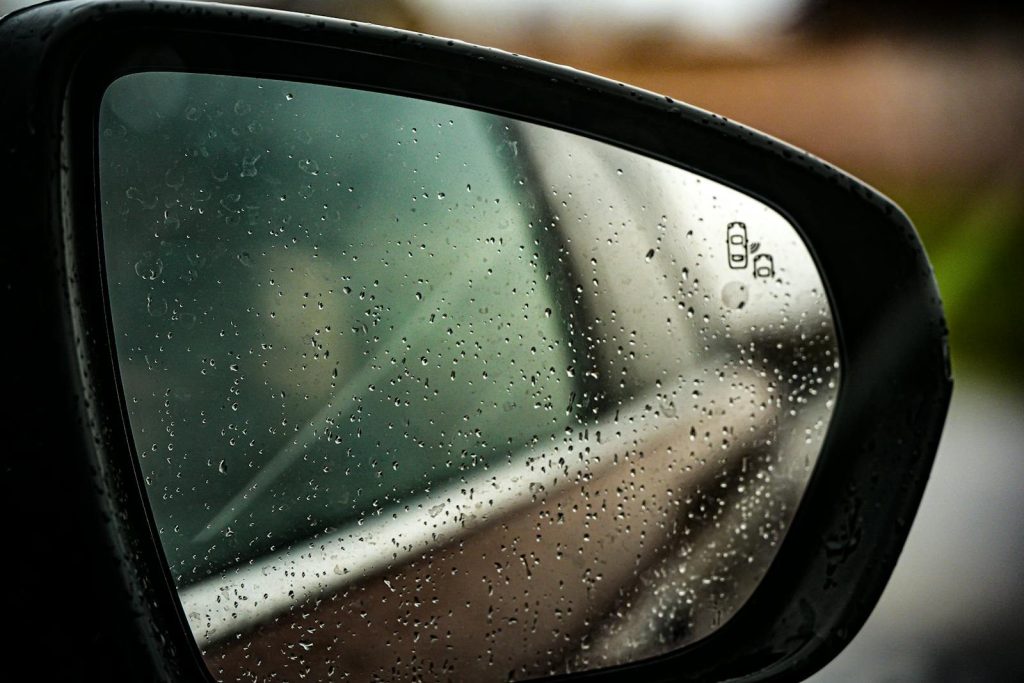 Close-up of a car side mirror with raindrops, reflecting a blurred background.