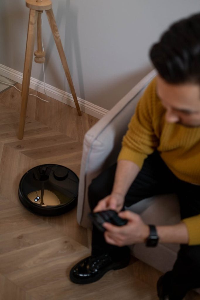 Adult male using remote control to manage robotic vacuum cleaner on wooden flooring indoors.