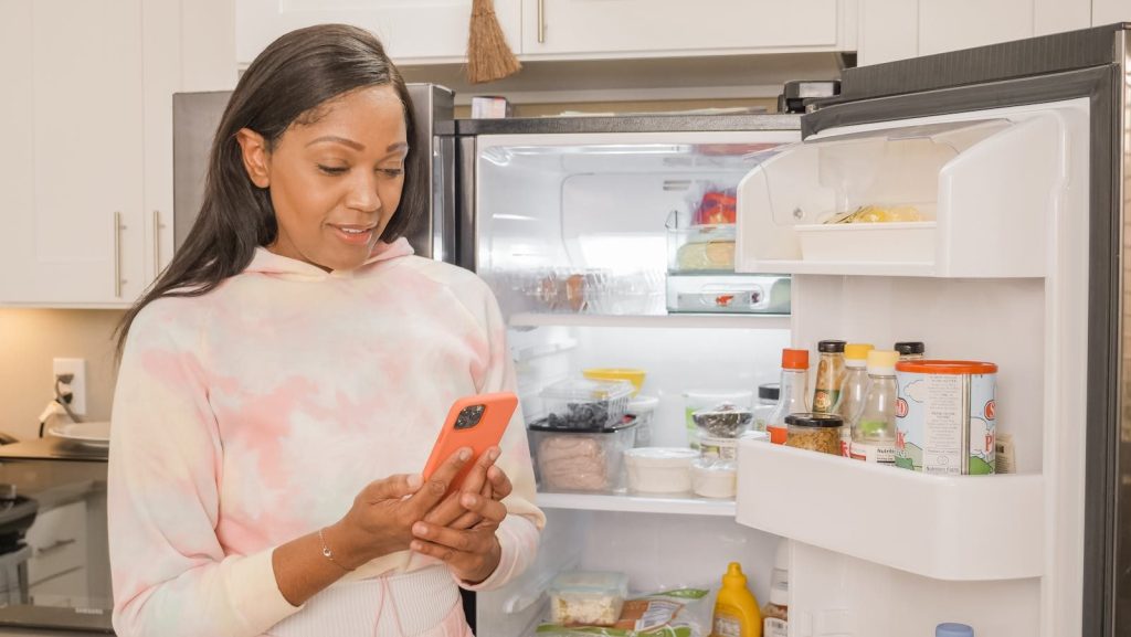 Woman using smartphone for online shopping while standing in the kitchen with an open refrigerator.