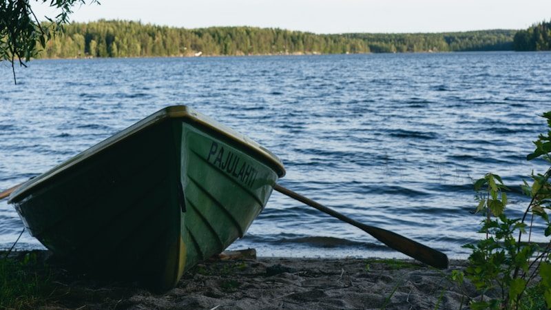 Green rowboat rests on a sandy shore near lake.