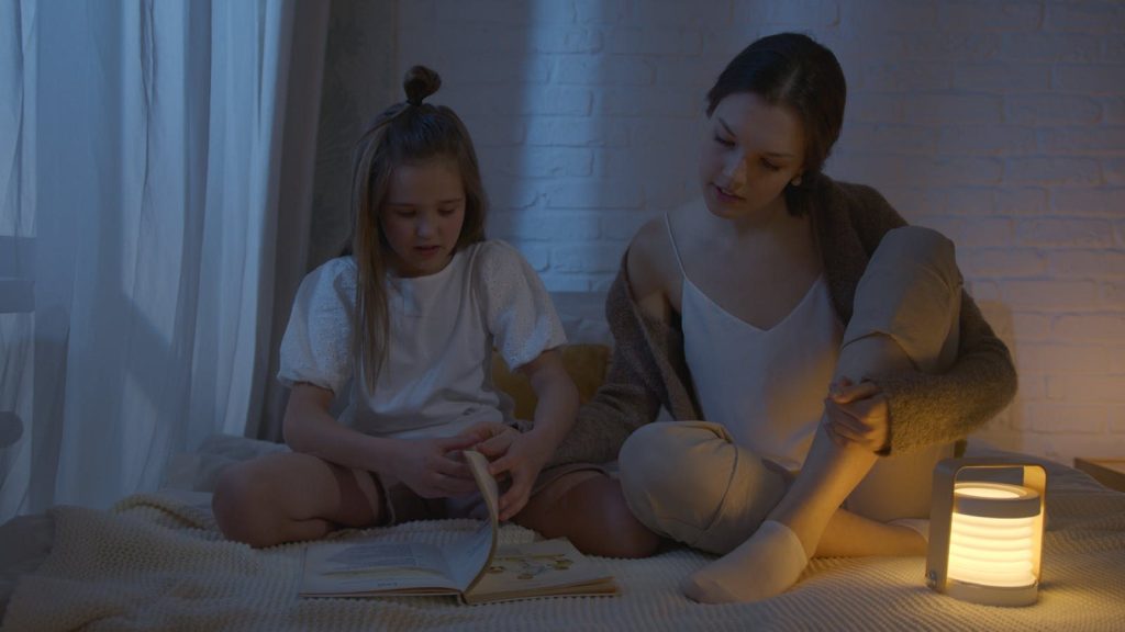 A mother and daughter bond over a book, creating a cozy night-time reading scene with soft lighting.