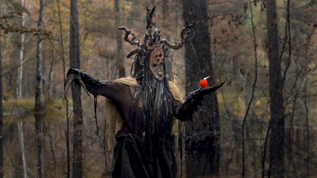 Eerie figure with mask and red candle in a haunting forest setting.