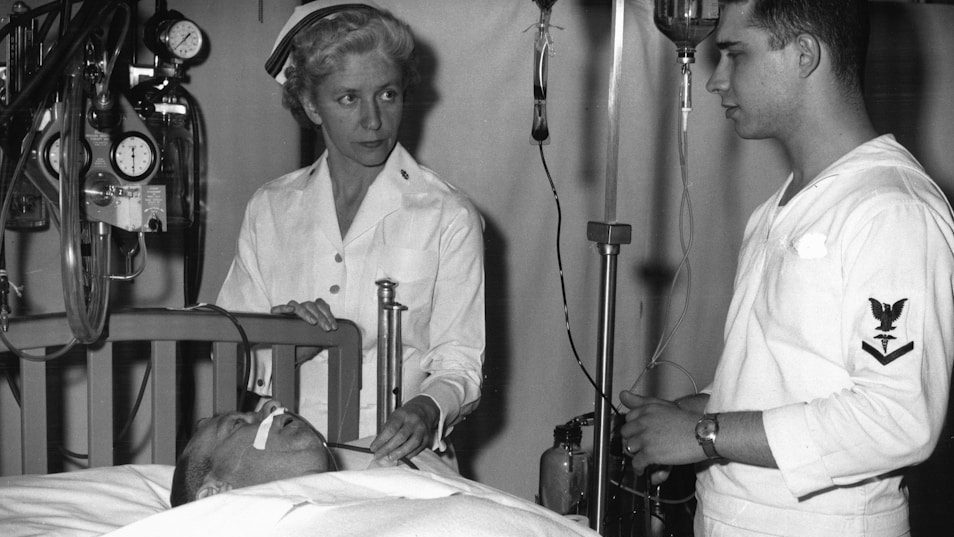 Nurse and sailor attend patient in hospital bed.