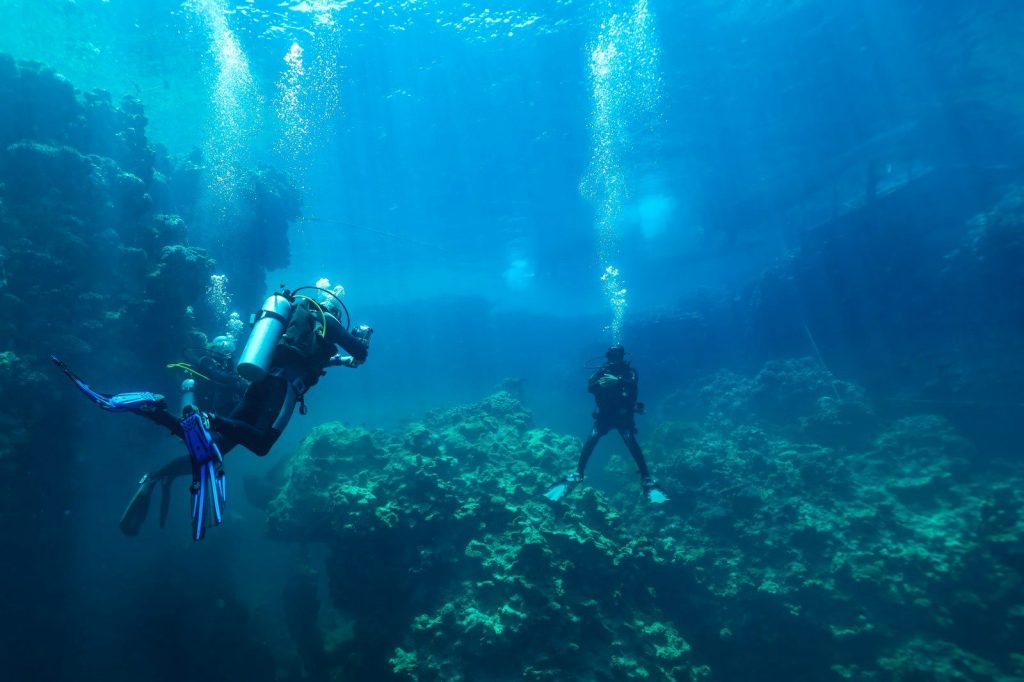 Divers explore a coral reef underwater with bubbles rising.