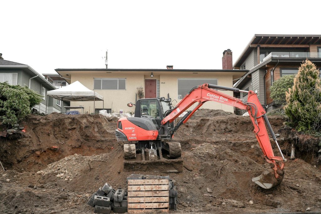 a red excavator digging a pile of dirt in front of a house