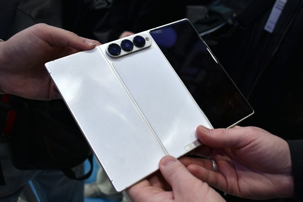 Hands hold a white foldable smartphone with three cameras.