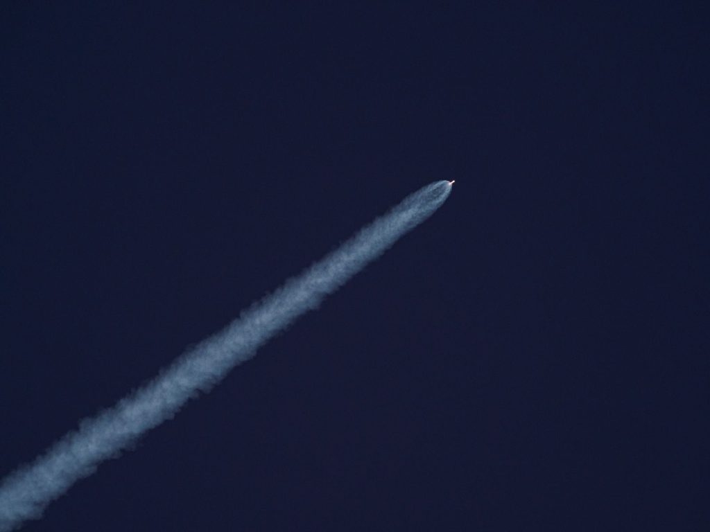 A rocket ascends into the sky, leaving a trail of smoke against the clear blue sky.
