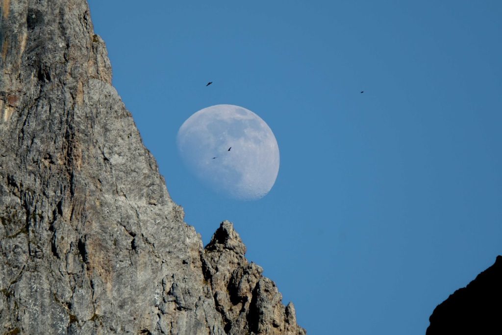 Stunning view of the moon rising above jagged mountain formations under a clear blue sky.