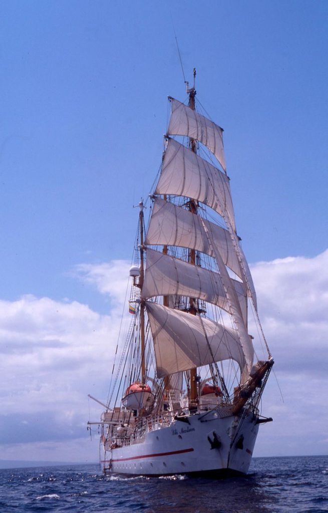 A classic tall ship with full sails glides over the ocean against a backdrop of blue sky and clouds.