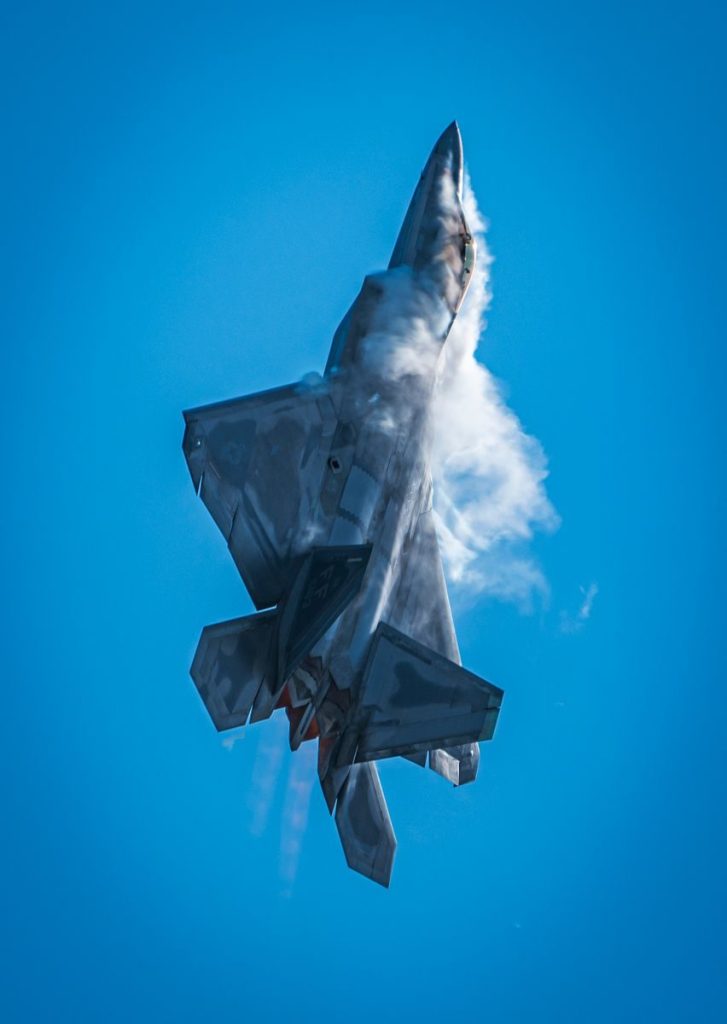 A fighter jet performs a vertical maneuver with vapor trails against a clear blue sky.