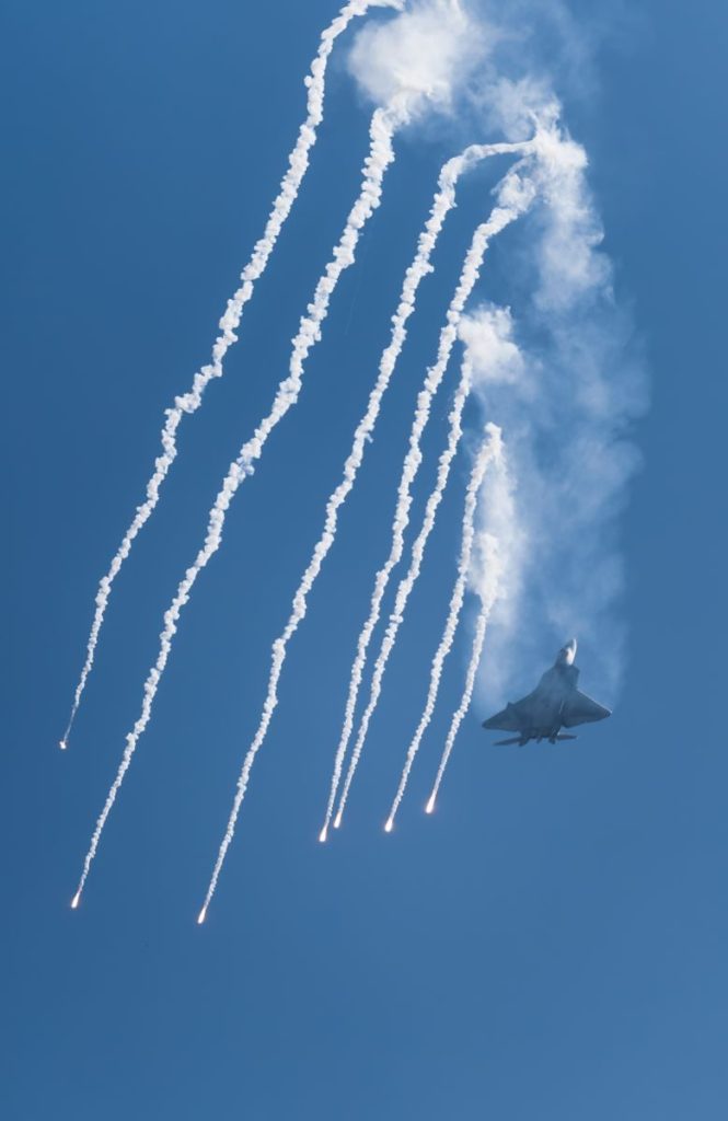 F-22 Raptor jet releasing flares in a dynamic air show display against a clear blue sky.