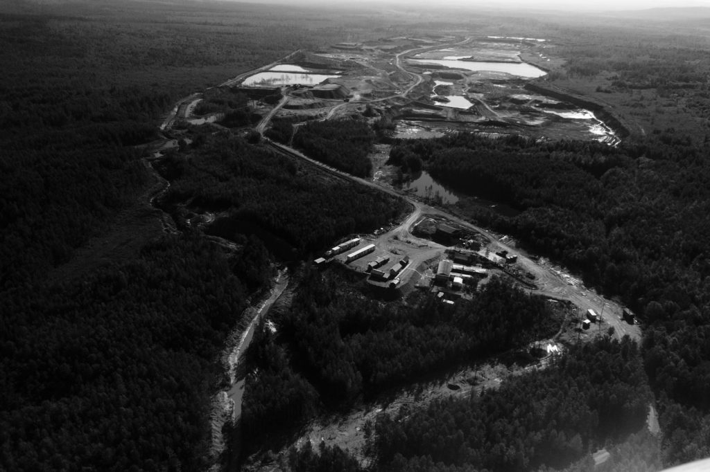 A black and white aerial photograph of an industrial site nestled within a lush forest landscape.