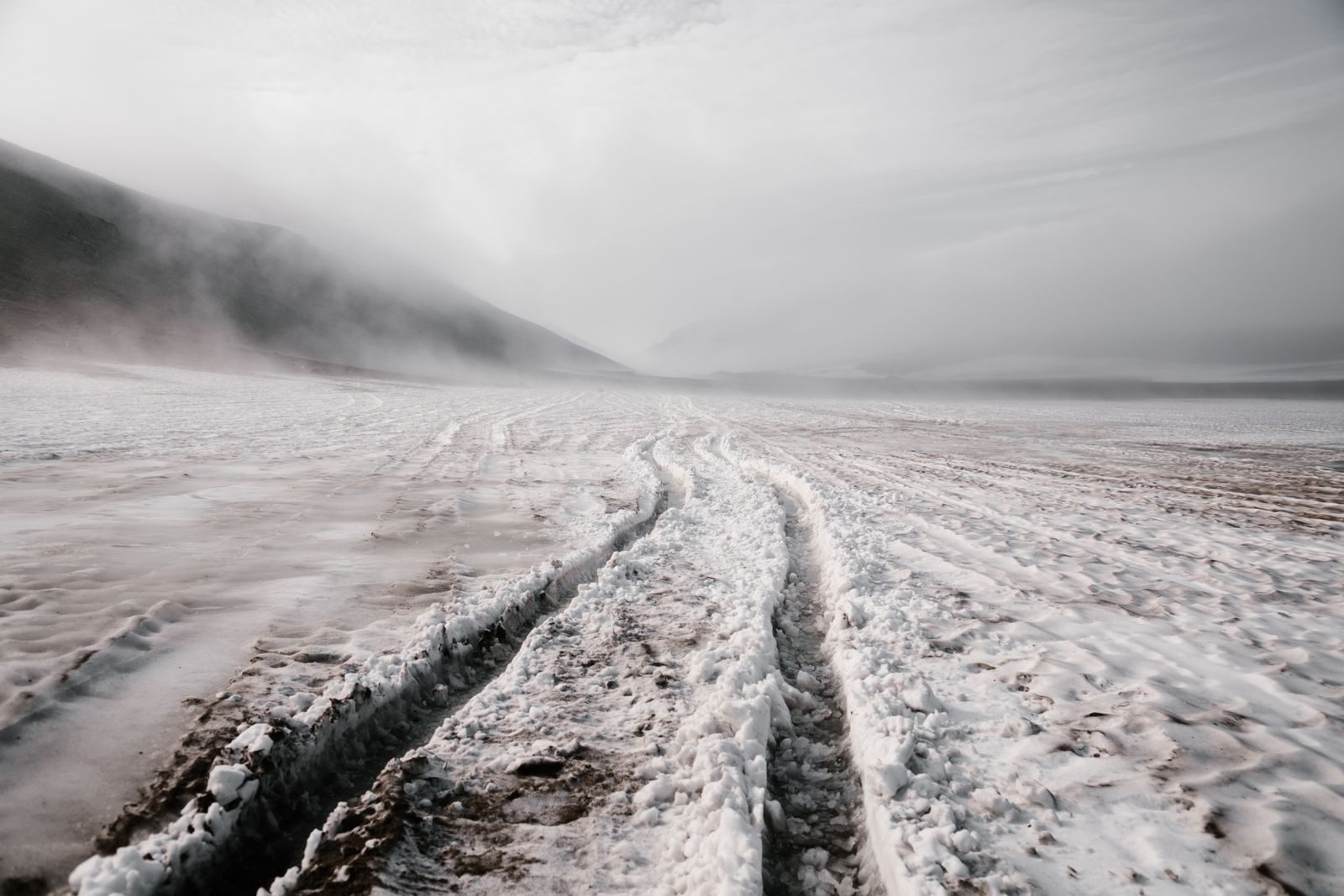A foggy, snow-covered landscape with visible tire tracks creating patterns.