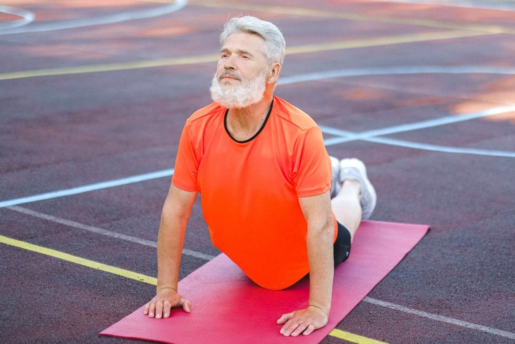 Full length of determined elderly male with stylish hairstyle and beard stretching on fitness mat while training alone