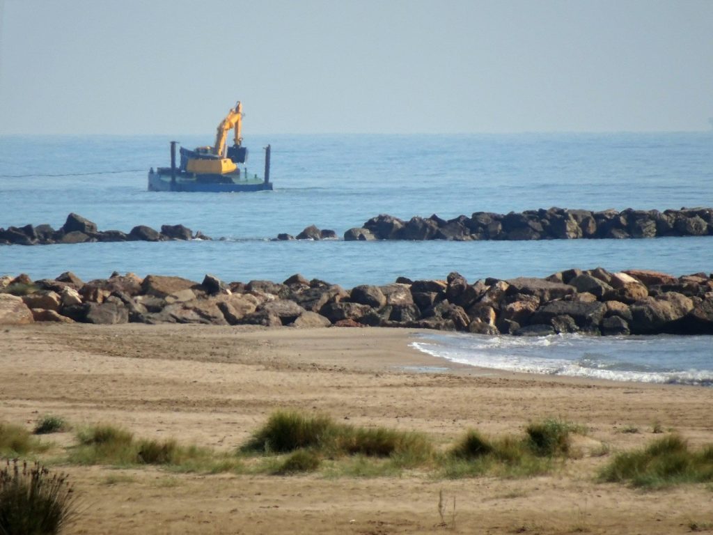 Excavator on barge working in the sea near shore.