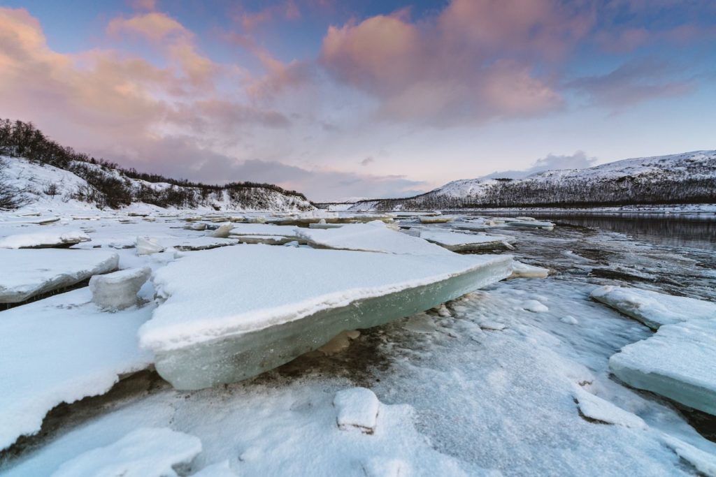 Serene frozen lake landscape surrounded by snow-covered mountains and icy waters at sunset.