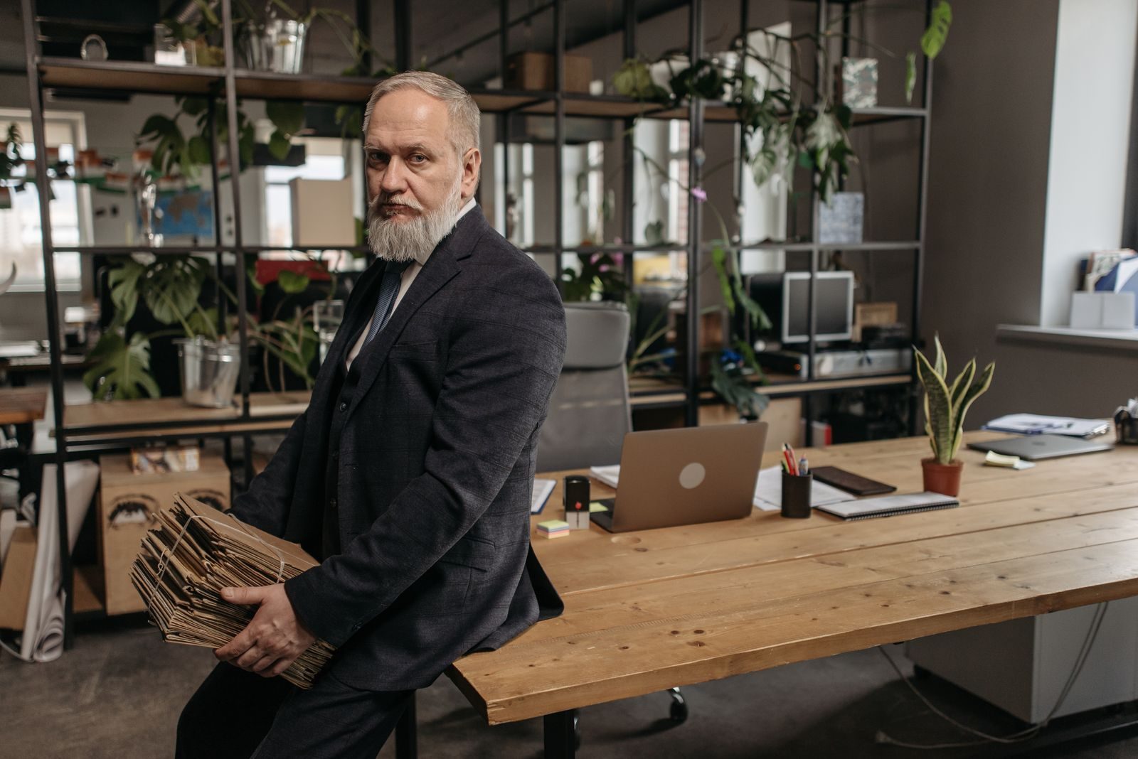 Elderly businessman in a dark suit holding folders in a modern office setting.
