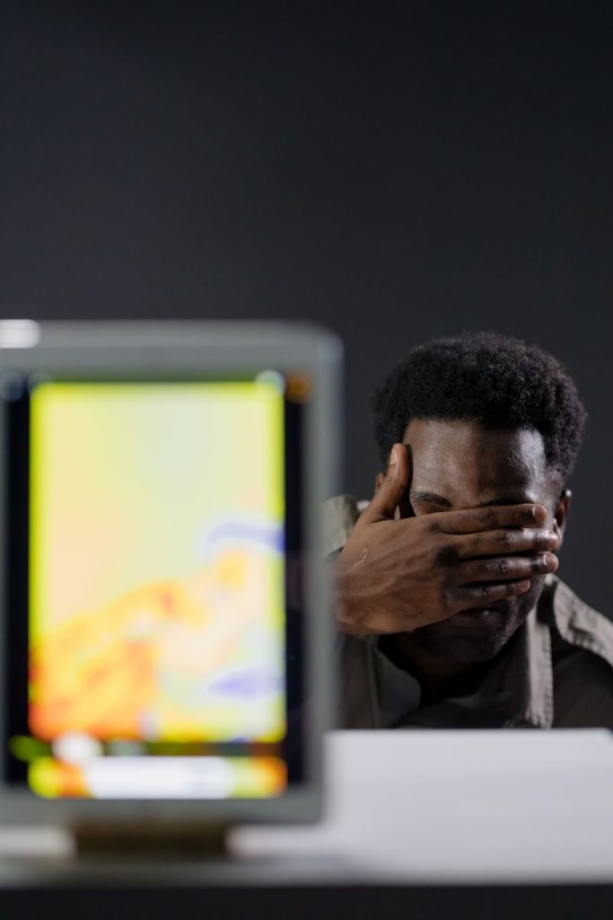 A man with short hair covers his face, seated behind a thermal imaging screen indoors.