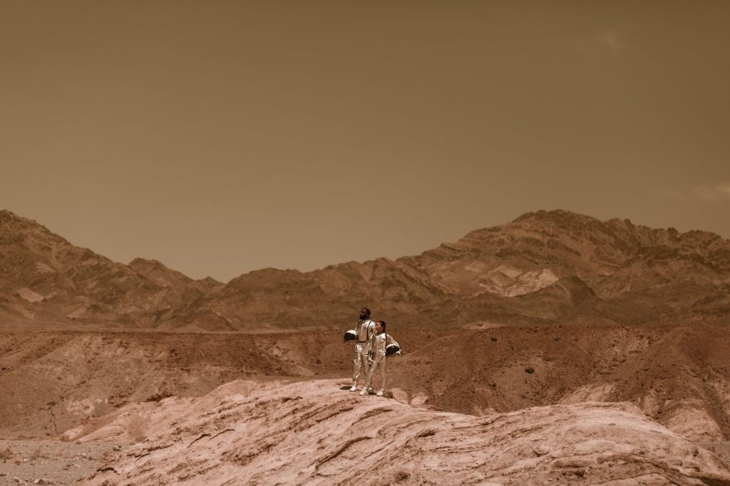 Two astronauts in space suits explore a Mars-like rocky desert landscape under a warm, sepia sky.