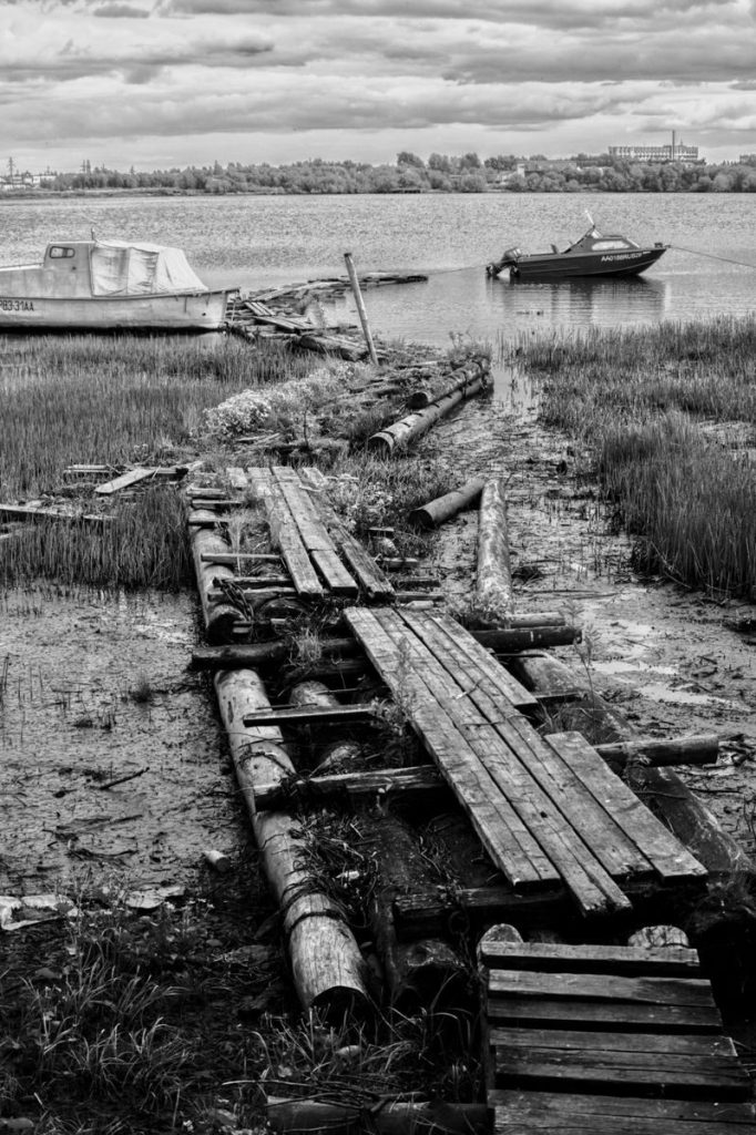 Weathered wooden deck leading to boats on a calm sea under overcast skies, in black and white.