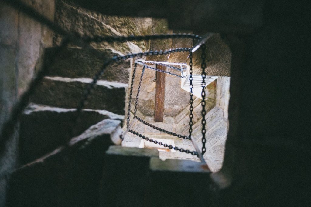 Looking up a spiral stone staircase with chains in a dimly lit setting, evoking mystery.