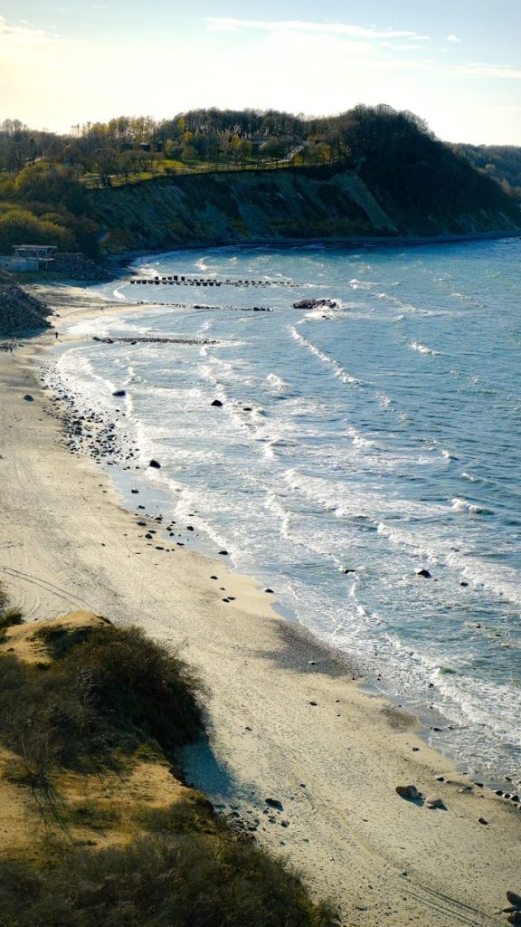 Sandy beach with gentle waves and a tree-covered cliff.