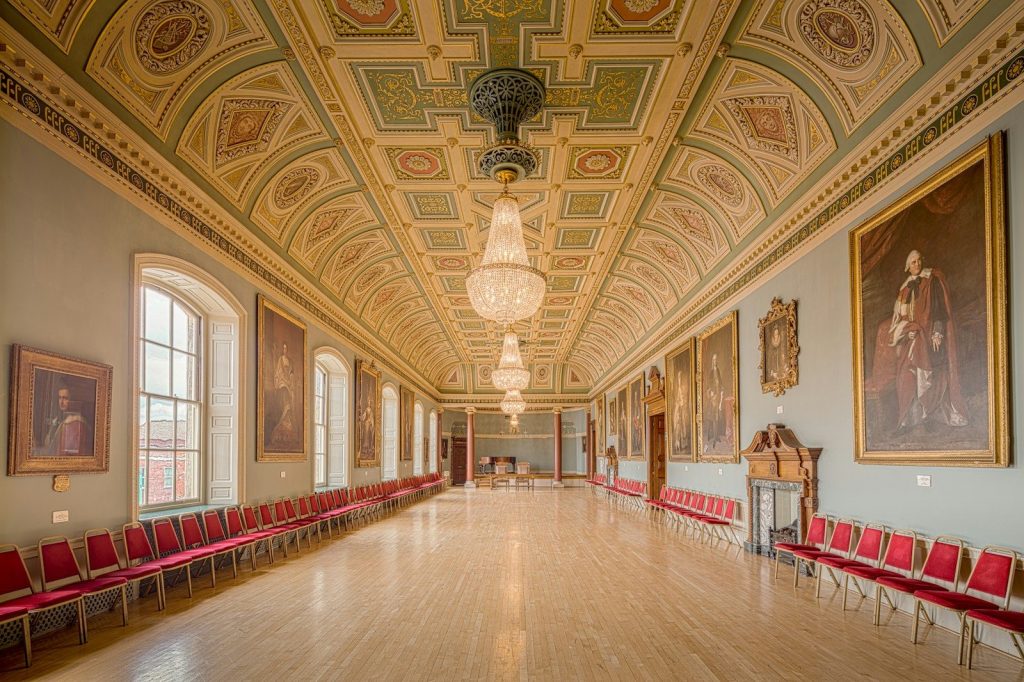 A long, ornate hall with chandeliers, portraits, and red chairs.