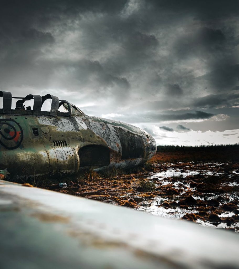 Rusty military aircraft wreckage in a muddy field under dramatic overcast skies, evoking a sense of history and desolation.