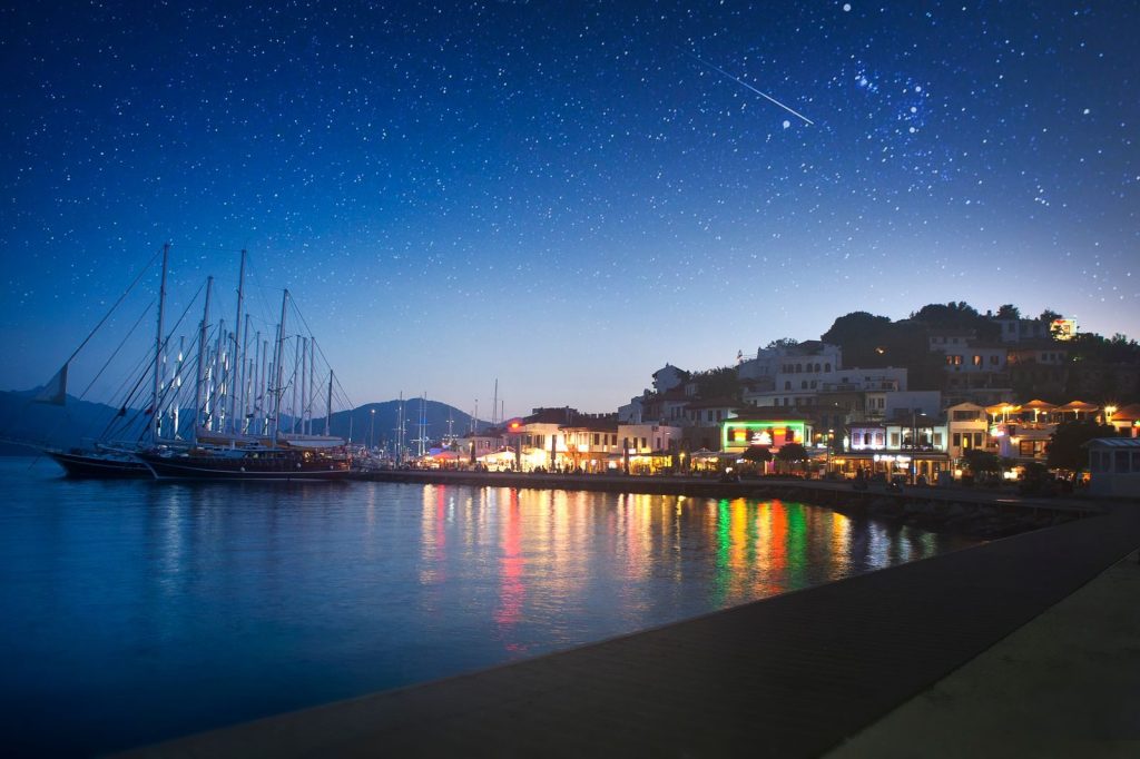 Nighttime view of a harbor with yachts and vibrant reflections under a starlit sky.