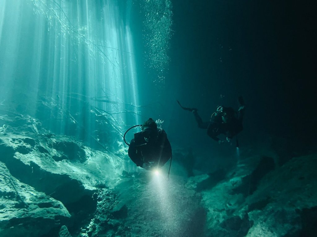 Scuba divers navigate underwater, exploring with flashlights and enjoying sunlight rays.
