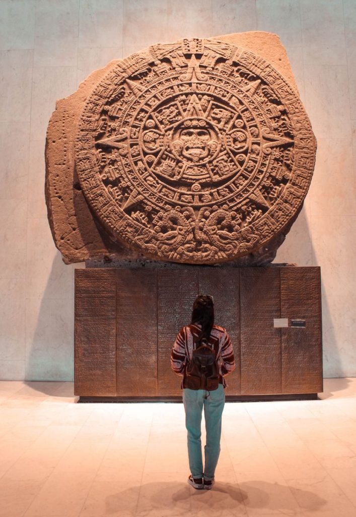 A woman observes the ancient Aztec Sun Stone in a museum setting, showcasing cultural heritage.