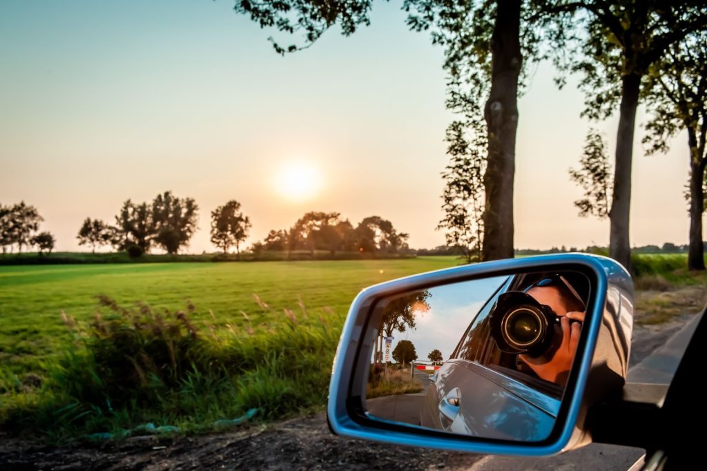 Photographer capturing a rural landscape in a car mirror at sunrise.