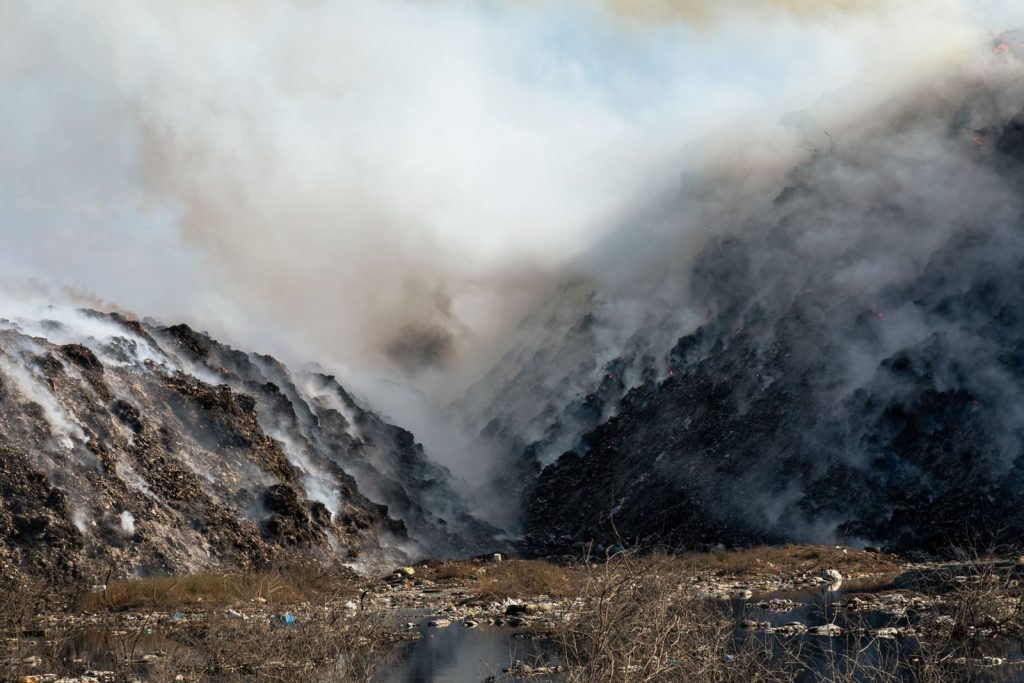 Thick smoke billows from a burning landfill surrounded by piles of waste and trash.
