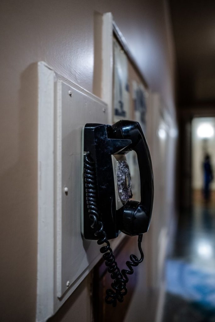 A classic wall-mounted black rotary dial telephone in a dimly lit hallway.