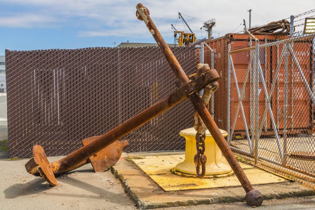A large rusty anchor alongside containers in a bustling shipyard.
