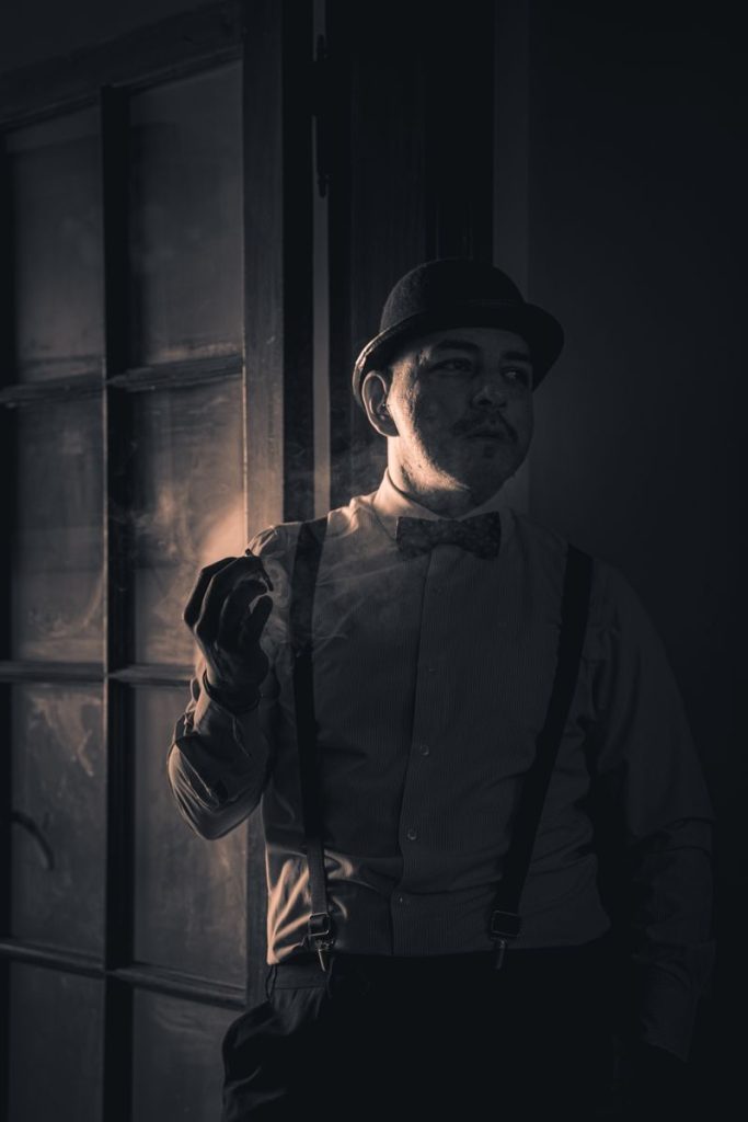 A vintage-style portrait of a man in a fedora and bow tie, smoking indoors with dramatic lighting.