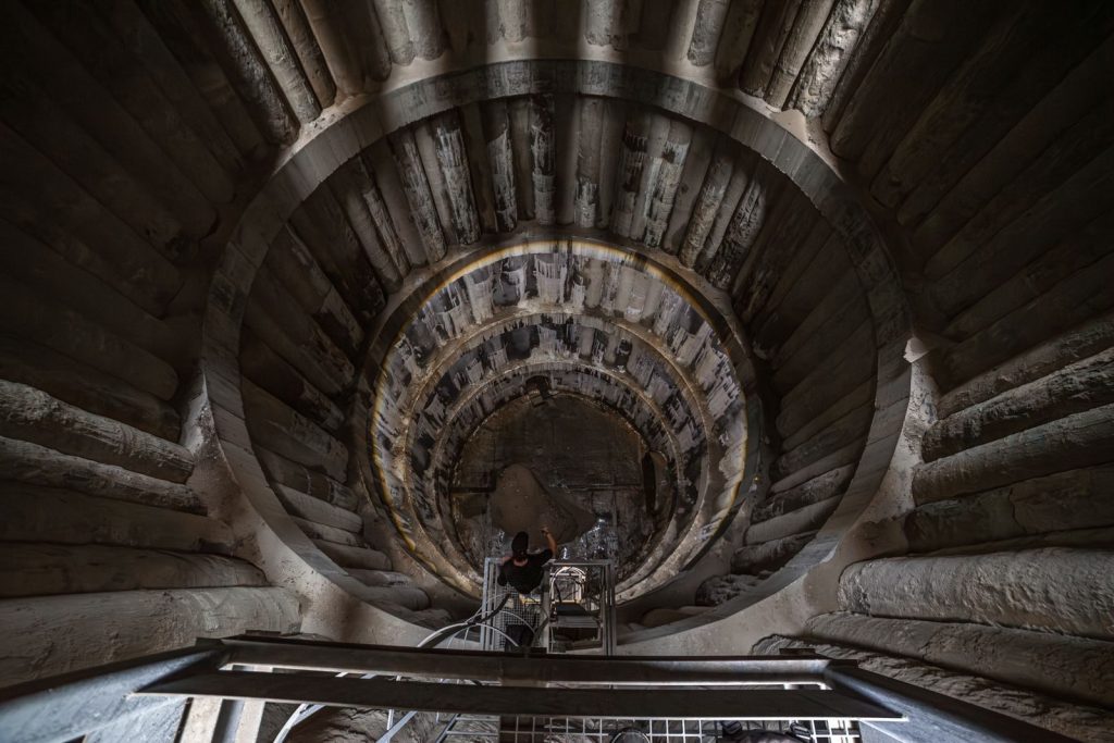 A worker stands inside a large, circular industrial tunnel, captured from above, showcasing engineering scale.