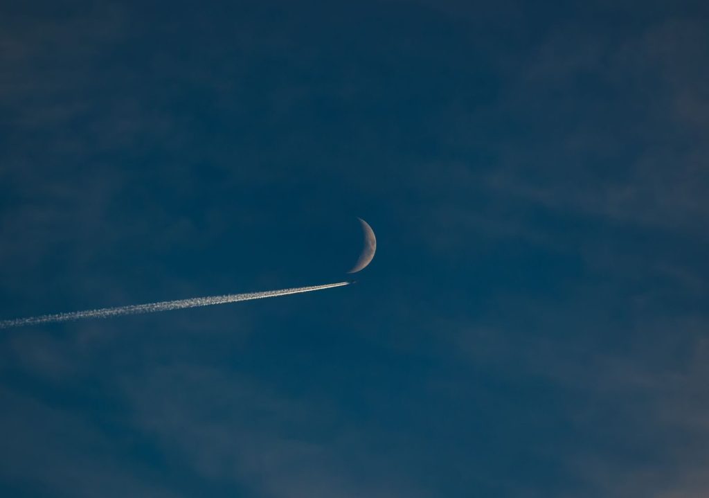 A stunning evening sky features a crescent moon and a jet contrail, creating a serene and dynamic scene.