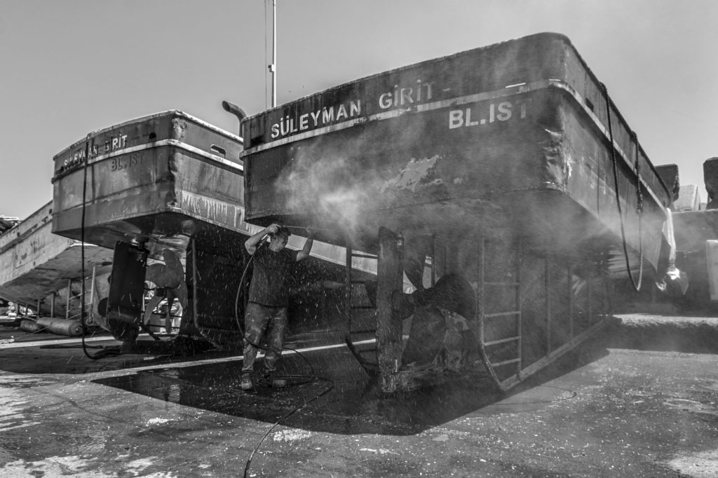 A worker cleaning boats at a shipyard with water spray in a black and white setting.