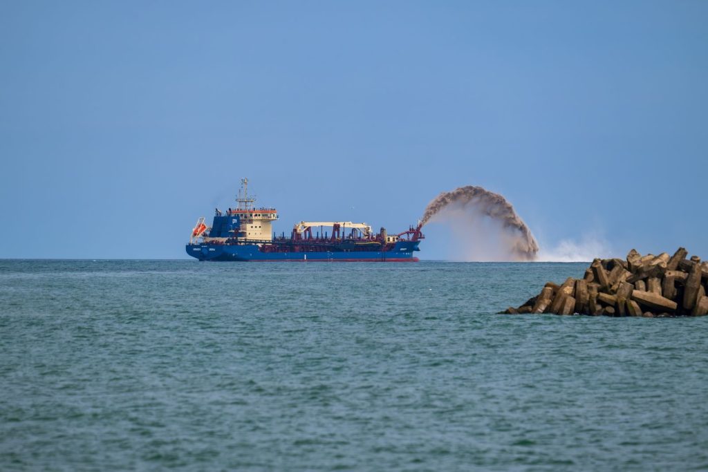 A blue dredger ship in the ocean carrying out dredging operations, visible with a dynamic sand spray.