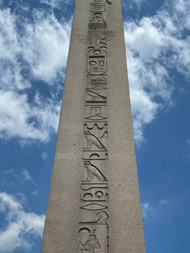 An ancient obelisk featuring intricate hieroglyphs, set against a bright blue sky with white clouds.