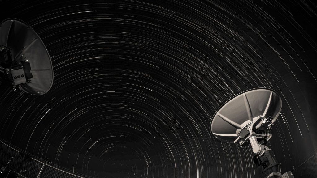 Long exposure of star trails creating circular patterns over two satellite dishes against a night sky.