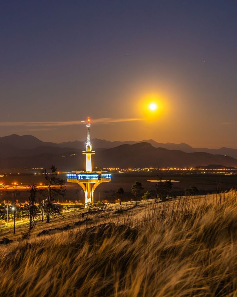 Television tower with artificial lights behind ridge under sky with bright sun in twilight