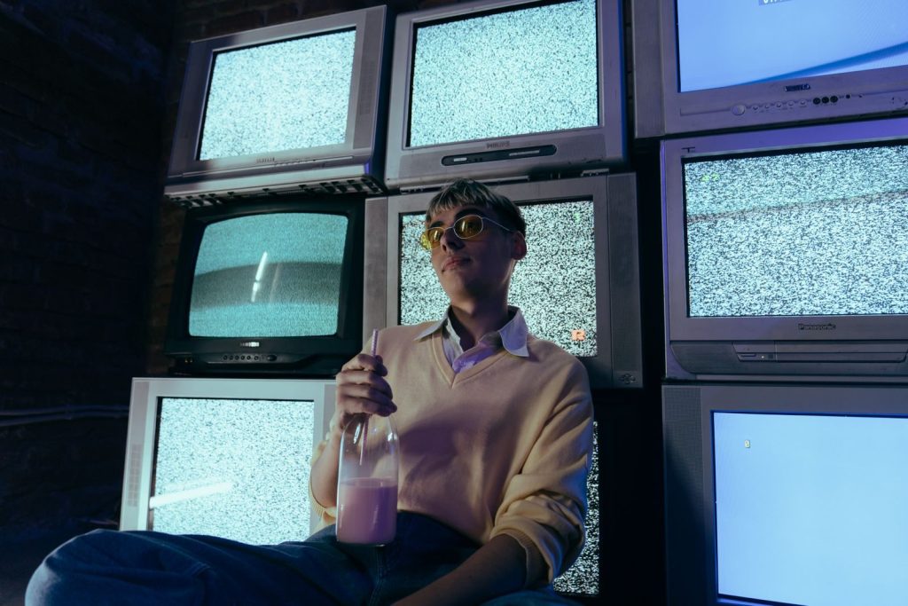 Teenage boy holding a milkshake surrounded by vintage CRT televisions showing static in a dimly lit room.